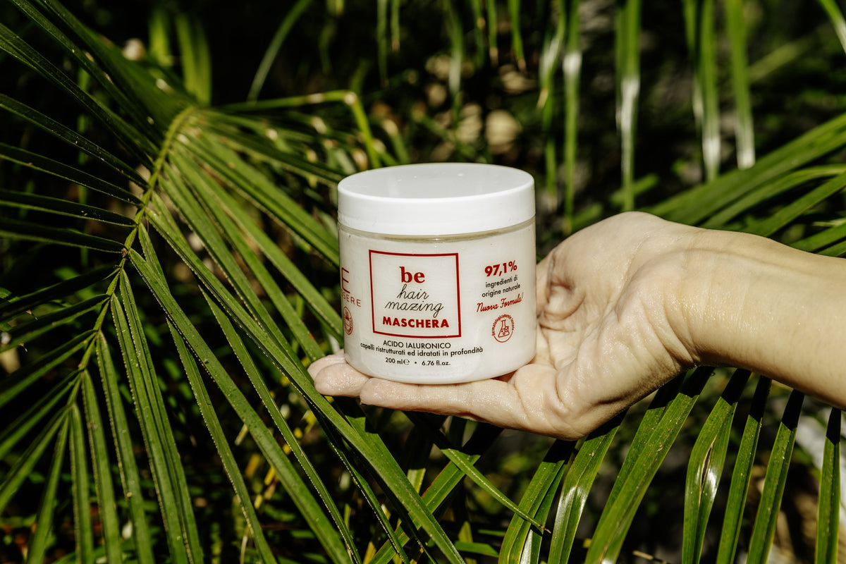 Una mano regge la maschera capelli Be Hair Mazing di Essere fra le foglie di palma, in pieno sole