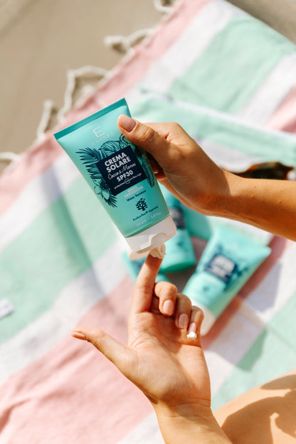 Crema Solare SPF 30 Essere Bio tenuta in mano sopra un telo a righe da spiaggia, gesto d'uso al sole
