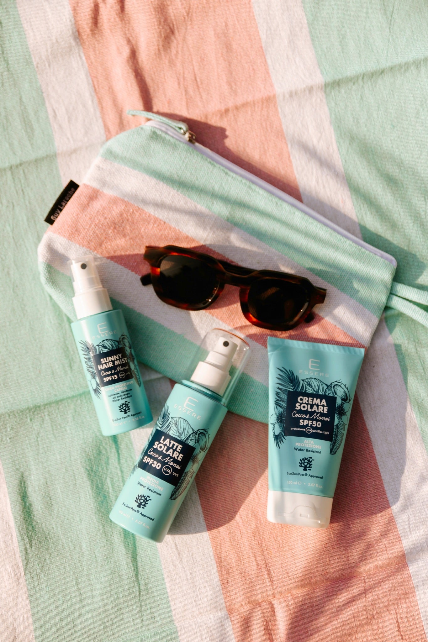 Solari Be Sunny Essere Bio in flat-lay su telo da spiaggia con occhiali e pochette, crema e spray SPF