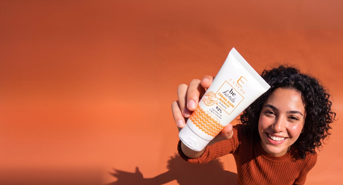 Ragazza dai capelli ricci con un maglione a coste ruggine, ripresa dall'alto su un fondo terracotta, sorride e tende verso l'obiettivo il tubetto di Crema Mani Mango Be Hands di Essere